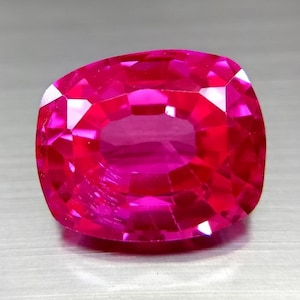 Puede incluir: Una gema de rubí tallada en cojín. La gema es de un rojo vibrante y saturado, con un alto grado de claridad. Las facetas reflejan la luz, creando una exhibición brillante. La gema se presenta sobre un fondo neutro.