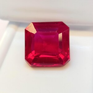 May include: A faceted, square-cut ruby gemstone. The gem is a deep, vibrant red color with a high degree of clarity. The gemstone is set against a white background, highlighting its brilliance and color. The cut of the stone allows for maximum light reflection.