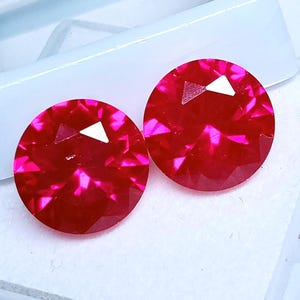 May include: Two round, faceted, bright red gemstones with a triangular white facet. The gems are displayed in a white plastic case. The gems have a brilliant, reflective surface.