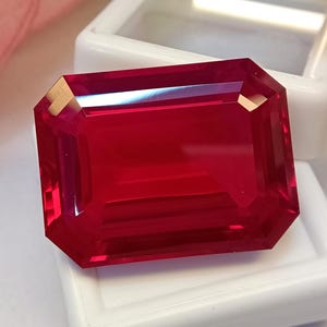 Könnte beinhalten: Ein leuchtend roter Edelstein im Rechteckschliff mit einer facettierten Oberfläche, die das Licht reflektiert. Der Stein liegt auf einem weißen Präsentationssockel und zeigt seine tiefrote Farbe sowie eine klare, transparente Beschaffenheit.