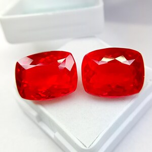 May include: Two faceted, cushion-cut, bright red gemstones. The gems are displayed on a white surface inside a white container. The gems have a high degree of clarity and brilliance, reflecting light.