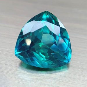 May include: A faceted, triangular gemstone with a vibrant teal color. The gem's surface reflects light, showcasing its intricate cuts and angles. The gemstone is set against a neutral, textured background.