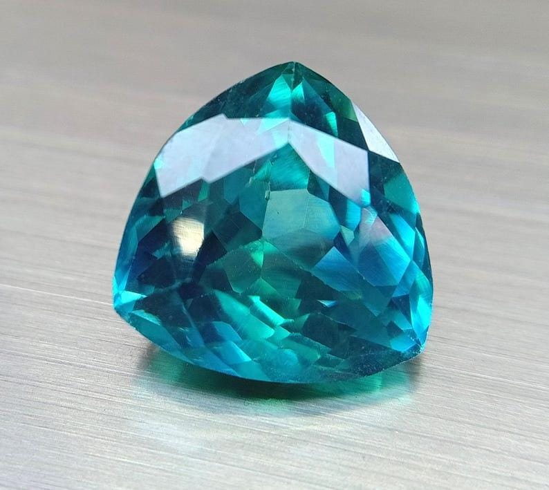May include: A faceted, triangular gemstone with a vibrant teal color. The gem's surface reflects light, showcasing its intricate cuts and angles. The gemstone is set against a neutral, textured background.