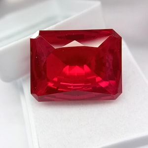 May include: A faceted, rectangular-cut, deep red gemstone. The gem is displayed in a white box, showcasing its brilliant, reflective surfaces. The stone's facets catch and reflect light, creating a vibrant display of color and brilliance.