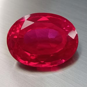 Pode incluir: Uma gema de rubi facetada em forma oval. A gema é de cor vermelha profunda e saturada, com alto grau de clareza. As facetas refletem a luz, criando uma aparência brilhante. O rubi está sobre um fundo neutro e claro.