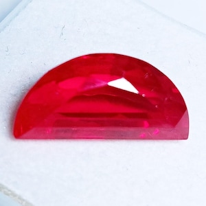 May include: A faceted, half-moon shaped, vibrant red gemstone. The gem has a flat base and a curved top, with multiple facets that catch the light. The gemstone is set against a white background.