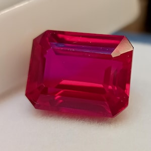 Peut inclure: Une pierre précieuse d'un rouge rosé profond et vibrant, taillée en forme d'émeraude rectangulaire. La pierre présente des facettes nettes et précises qui reflètent la lumière, offrant un aspect translucide avec une surface polie.