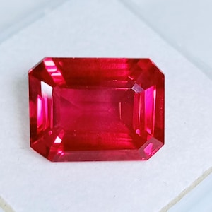 May include: A faceted, rectangular-cut ruby gemstone. The gem is a deep, vibrant red color with sharp, clean edges and a polished surface. The gemstone is set against a white background, highlighting its brilliance.
