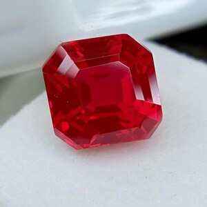 May include: A faceted, radiant-cut, red gemstone. The gem has a square shape with beveled edges, reflecting light and creating a brilliant display of color. The gemstone is set against a white background, highlighting its vibrant hue.