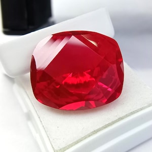 May include: A vibrant red, cushion-cut gemstone with multiple facets. The gem is presented in a white box, highlighting its cut and colour. The facets reflect light, creating a sparkling effect. The overall presentation suggests a high-quality precious stone.