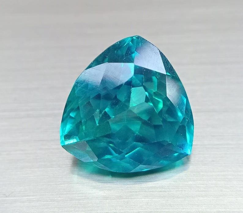 May include: A faceted, triangular gemstone with a vibrant teal color. The gem's surface reflects light, showcasing its intricate cuts and angles. The gemstone is a striking example of fine jewelry.