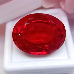 Könnte beinhalten: Ein facettierter, ovalförmiger, tiefroter Edelstein. Der Edelstein liegt vor einem weißen Hintergrund, mit weichen rosa und weißen Elementen im Hintergrund. Die Facetten des Edelsteins reflektieren das Licht.