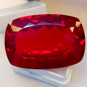 Può includere: Una grande gemma sfaccettata, taglio a cuscino, di colore rosso intenso. La gemma è esposta in una scatola bianca, che ne esalta la brillantezza e la chiarezza. Il taglio crea molteplici sfaccettature.