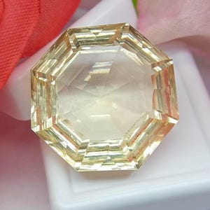 May include: A faceted, octagonal gemstone with a pale yellow hue. The gem is set against a white background, with red and pink fabric in the background. The gem's facets reflect light, creating a sparkling effect.