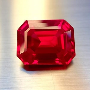 May include: A vibrant, faceted, emerald-cut ruby gemstone. The deep red stone has a rectangular shape with beveled edges, reflecting light and showcasing its clarity. The gemstone is set against a neutral background.