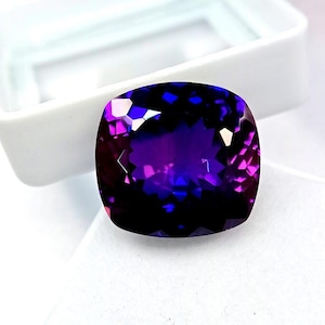 May include: A large, faceted gemstone with a cushion cut. The gem displays a deep purple hue with flashes of blue and pink, set against a white background. The stone is likely a high-quality amethyst or similar precious stone.
