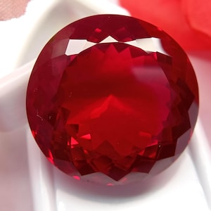 May include: A large, round, faceted, deep red gemstone. The gem is set against a white background, with a hint of pink and red in the background. The gemstone's many facets reflect light, creating a brilliant display.