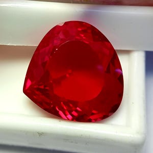 Puede incluir: Una gema de color rojo intenso y vibrante con un corte triangular facetado. La piedra presenta una superficie pulida con numerosas facetas geométricas que reflejan la luz, creando un aspecto brillante. La gema se encuentra sobre un fondo blanco.