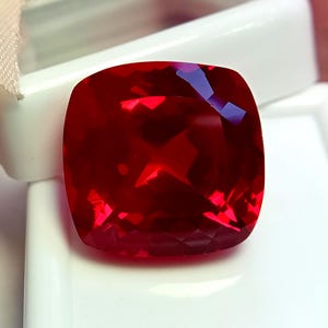 May include: A vibrant, cushion-cut red gemstone with a faceted surface that reflects light. The stone features a square shape with rounded corners and a deep, saturated crimson hue, displaying clear internal reflections across its polished facets.
