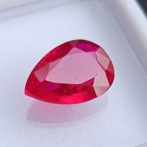 Peut inclure: Une pierre précieuse en rubis en forme de poire et à facettes. La gemme est d'un rouge vif avec de multiples facettes qui réfléchissent la lumière. La pierre précieuse est posée sur un fond blanc.