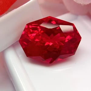 May include: A faceted, octagonal-cut, deep red gemstone. The gem has many reflective surfaces, catching the light and creating a brilliant display. The gemstone is set against a white background.
