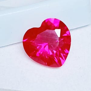 Può includere: Una gemma sfaccettata a forma di cuore, in una vibrante tonalità rosso-rosa. La gemma ha molteplici sfaccettature che riflettono la luce, creando un effetto scintillante. La forma a cuore è ben definita, con una parte superiore arrotondata e una inferiore a punta.