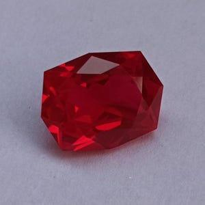 Peut inclure: Une pierre précieuse rouge vif, taillée à facettes, de forme géométrique. La gemme présente de multiples surfaces réfléchissantes, créant un jeu de lumière et de couleur. La pierre est posée sur un fond clair et uni.
