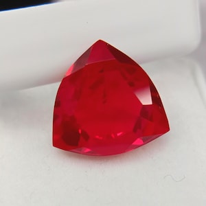 May include: A faceted, triangular-shaped, vibrant red gemstone. The gem is set against a white background, showcasing its brilliant cut and intense color. The gemstone's facets reflect light, creating a sparkling effect.