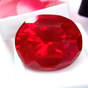 Peut inclure: Une pierre précieuse rouge foncé, de forme ovale et à facettes. La gemme présente une surface brillante et réfléchissante, avec de multiples facettes qui captent et réfléchissent la lumière. La pierre est posée sur un fond blanc, soulignant sa couleur vive et sa clarté. Cette pierre est adaptée à la joaillerie.