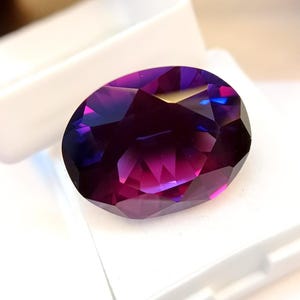 Puede incluir: Una gema ovalada con un corte facetado, que muestra un degradado de colores desde el púrpura intenso hasta el magenta vibrante y toques de azul. La gema se encuentra sobre un fondo blanco, resaltando su brillo y claridad. Esta gema podría usarse para joyería.
