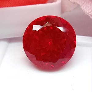 May include: A faceted, round, deep red gemstone. The gem is set against a white background, with a hint of red and pink fabric in the background. The gem's surface reflects light, showcasing its intricate cuts and vibrant color.