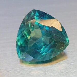 May include: A faceted, triangular gemstone with a gradient of teal and green hues. The gem's surface reflects light, showcasing its intricate cuts and internal structure. The gemstone is a striking example of fine jewelry.