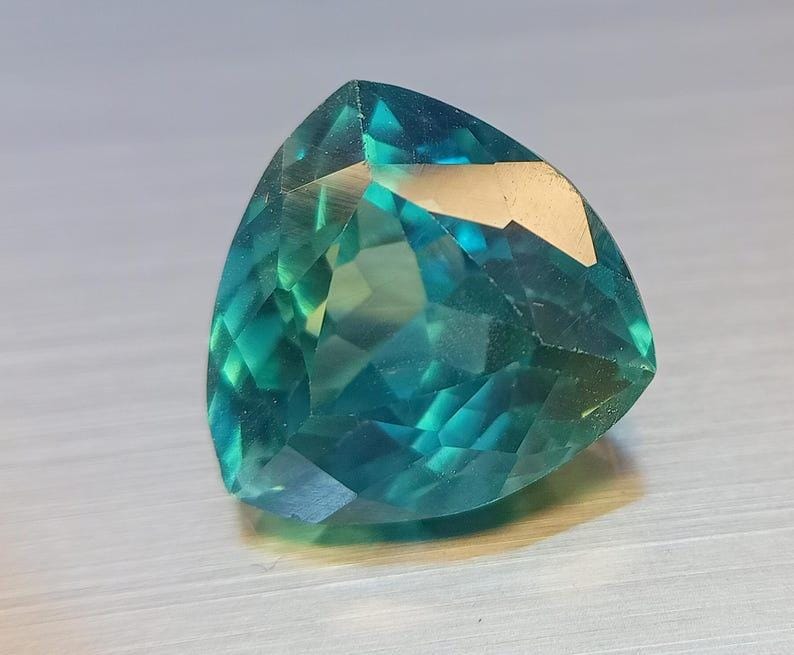 May include: A faceted, triangular gemstone with a gradient of teal and green hues. The gem's surface reflects light, showcasing its intricate cuts and internal structure. The gemstone is a striking example of fine jewelry.