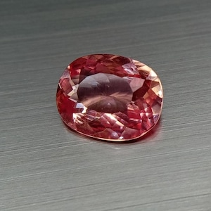 Op de afbeelding: Een gefacetteerde, ovaalvormige edelsteen met een perzikroze tint. De edelsteen vertoont interne reflecties en een gepolijst oppervlak, tegen een grijze achtergrond. De slijpvorm van de edelsteen onthult meerdere facetten, waardoor de glans wordt versterkt.
