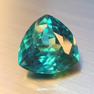 May include: A faceted, triangular gemstone with a teal and green color gradient. The gem has a polished surface with multiple facets that reflect light, creating a sparkling effect. The gemstone is set against a neutral background.