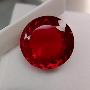 Puede incluir: Una gema grande, redonda y facetada, de color rojo intenso. La gema tiene un corte brillante, que refleja la luz y crea un efecto brillante. La gema está sobre un fondo blanco.