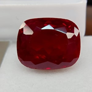 May include: A faceted, cushion-cut ruby gemstone. The gem is a deep, saturated red colour with multiple facets that reflect light. The shape is square with rounded corners, and the stone is set against a white background.