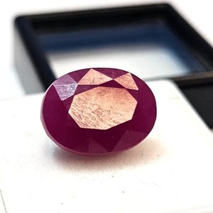 May include: Oval-cut ruby gemstone with a faceted surface. The gem is a deep red color with lighter internal patterns. The ruby is set against a white surface, with a dark blue and black background.