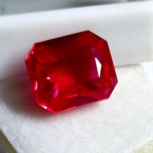 Może przedstawiać: Fasetowany, prostokątny kamień szlachetny rubinu. Klejnot ma żywy, głęboki czerwony kolor z ostrymi, zdefiniowanymi krawędziami i fasetami. Rubin jest umieszczony na białym tle.
