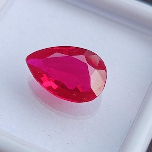 Peut inclure: Une pierre précieuse rubis en forme de poire et à facettes. La gemme est d'un rouge profond et vibrant, avec de multiples facettes captant la lumière. Le rubis est posé sur un fond blanc, soulignant son éclat et sa clarté. Un gros plan met en valeur la surface polie de la gemme.