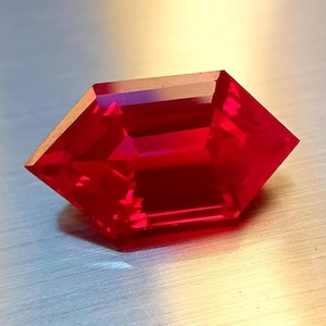 Puede incluir: Una gema roja facetada con una forma geométrica única. La gema tiene un color intenso y rico y múltiples facetas que reflejan la luz. El corte de la piedra es un hexágono modificado, con lados angulados y una parte superior plana.