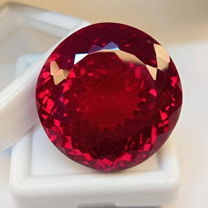 May include: A large, round, faceted, deep red gemstone. The gem is displayed against a white background, highlighting its brilliant cut and vibrant colour. The gemstone's facets reflect light, creating a sparkling effect.