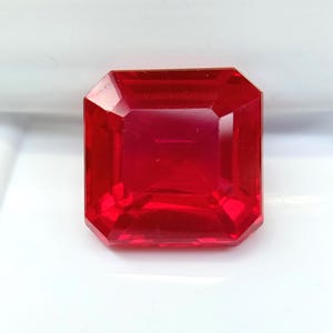 May include: A faceted, rectangular-cut, vibrant red gemstone. The gem has a high degree of clarity and brilliance, reflecting light across its many facets. The gemstone is set against a white background.