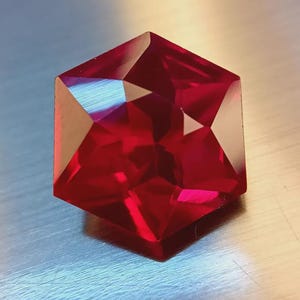 Op de afbeelding: Een gefacetteerde, zeshoekige edelsteen met een dieprode kleur. De vele facetten van de edelsteen reflecteren het licht, waardoor een briljante weergave van kleur en glans ontstaat. De edelsteen ligt op een metalen oppervlak.
