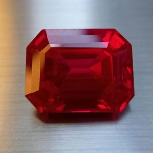 Puede incluir: Una gema roja oscura, tallada en forma rectangular y facetada. La gema tiene una superficie brillante y reflectante con múltiples facetas que capturan y reflejan la luz. La piedra preciosa se encuentra sobre un fondo metálico neutro.