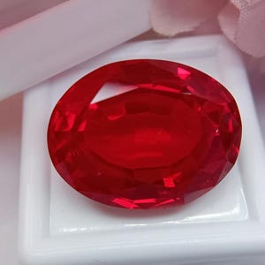 以下が含まれることがあります： 鮮やかな楕円形の赤い宝石。 宝石は白い背景に配置され、背景には柔らかいピンクと白のアクセントがあります。 宝石のファセットは光を反射し、鮮やかな色の表示を作成します。