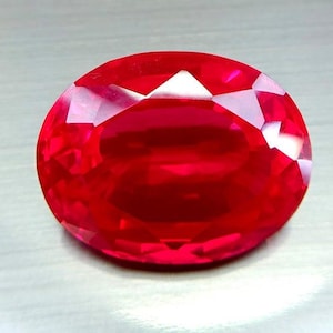 Puede incluir: Una gema roja vibrante, facetada y de forma ovalada. La superficie de la gema refleja la luz, mostrando sus intrincados cortes y brillo interno. La piedra preciosa es de un color rojo intenso y saturado, con una superficie pulida y reflectante.