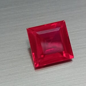 Op de afbeelding: Een vierkant geslepen, levendige rode edelsteen met een gefacetteerd oppervlak. De edelsteen is geplaatst tegen een geborstelde metalen achtergrond, die zijn briljante kleur en scherpe hoeken benadrukt. De vorm van de edelsteen is een perfect vierkant.