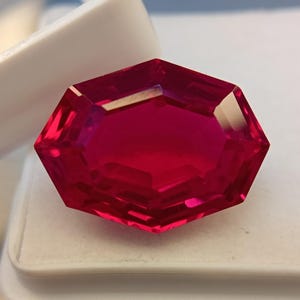 May include: A faceted, octagonal ruby gemstone. The gem is a deep, vibrant red color with multiple facets that reflect light. The ruby is set against a white background, highlighting its brilliance.