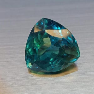 May include: A triangular-cut gemstone with facets, displaying shades of teal and green. The gem has a polished surface and reflects light, creating a sparkling effect. The gemstone is set against a neutral background.
